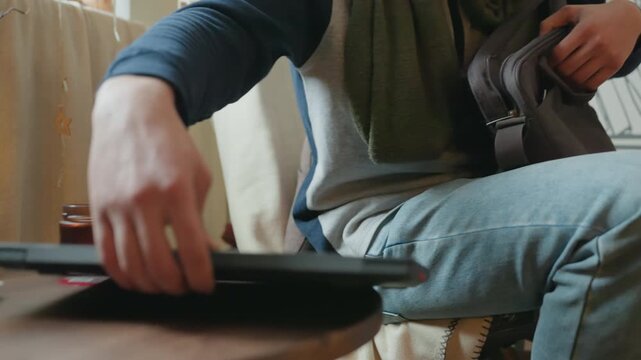 White man unzipping bag and placing laptop on lap in cozy livingroom, denim jeans and green scarf, closeup hands arranging device beside coffee on table, sequence shows packing, sliding laptop into