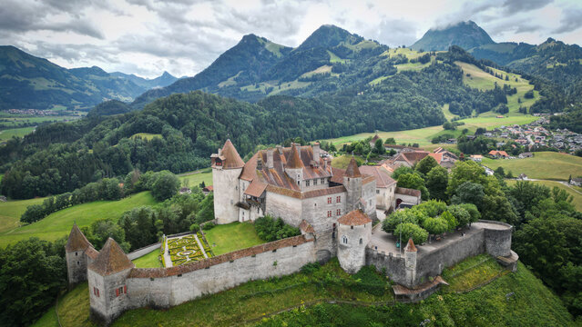 Aerial view of an ancient castle with red roofs and stone walls atop a green hill against a backdrop of majestic mountains, Gruyeres, Fribourg, Switzerland.