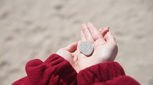 Hands holding smooth flat stones with red jacket and blurred sandy background in natural light