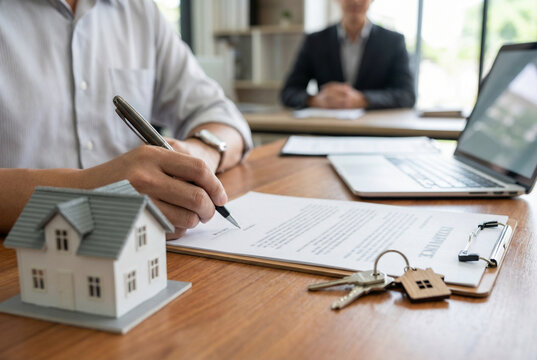 Close-up of person signing real estate agreement with house model and keys on desk, symbolizing property purchase, home ownership, contract finalization, and real estate transaction