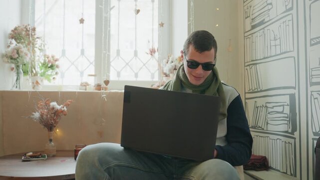 White man cozy window seat laptop, sunglasses and scarf, relaxed freelance designer working in sunlit studio, vintage decor with vases and illustrated bookshelf wallpaper, soft warm tones, focused
