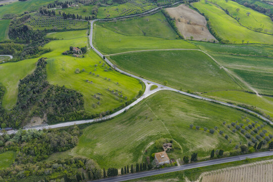 Aerial view of rolling green hills with winding roads, traditional farmhouses, and rows of cypress trees in the rural landscape of Pomarance, Tuscany, Italy.