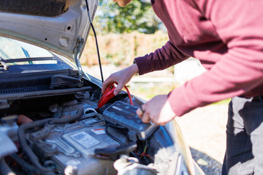 Young man using jumper cables to boost a car battery under the hood on a sunny day, roadside emergency assistance concept