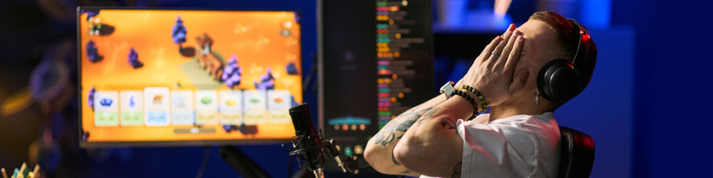 Young adult man streaming gameplay on computer, reacting with hands covering face while wearing headphones. Ideal for gaming content, live streaming, creator marketing, online entertainment