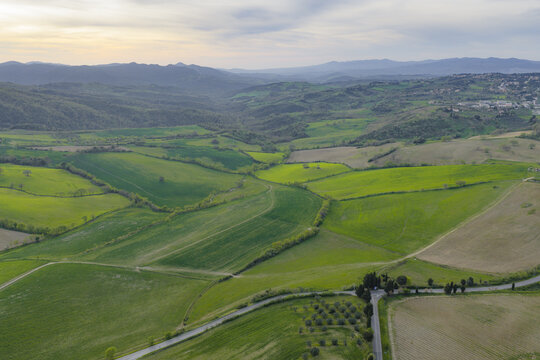 Aerial view of rolling green hills and agricultural fields with a winding road under a soft sunset sky in the countryside Pomarance, Tuscany, Italy.