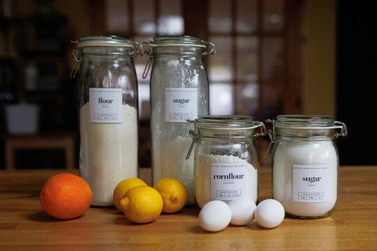 Baking ingredients including flour, sugar, cornflour, eggs, and fresh citrus fruits arranged on a wooden kitchen table