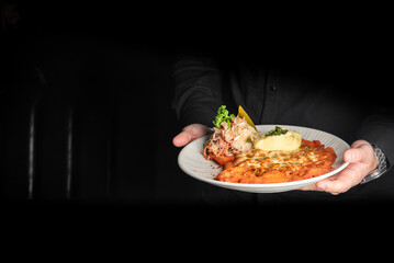A server's hands holding a white plate with a breaded schnitzel topped with melted cheese, served...