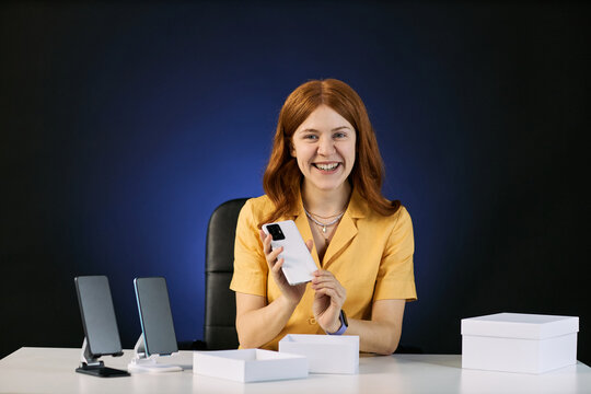 Portrait of young adult Caucasian woman holding smartphone and presenting product during livestream review. Useful for tech marketing, influencer promotion, ecommerce content, medium shot