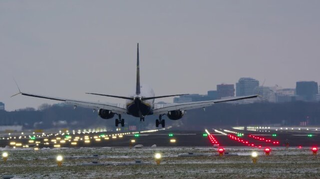 A commercial airplane approaches and lands on a brightly lit airport runway during an overcast winter evening. The camera captures the touchdown and rollout from a ground level view