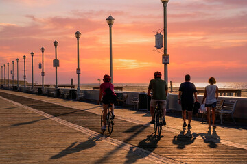 2 couples on the boardwalk at Ocean City MD during sunrise over the Atlantic Ocean.  Husband and wife are riding their bikes while another couple walks side by side.   Sunrise over the Ocean.   © Chet Wiker