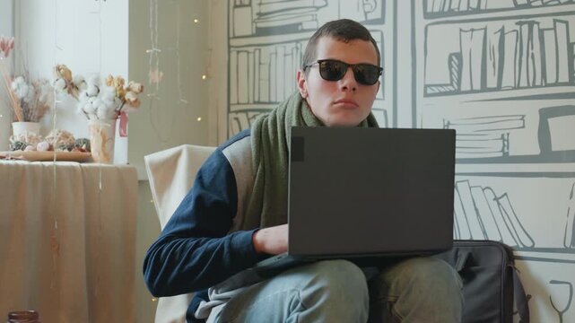 Caucasian student hunched over laptop, wearing sunglasses and green scarf, latenight study with weary expression, soft fairy lights and illustrated bookshelf mural, vase of dried flowers on table,