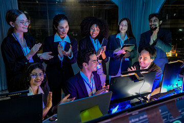 Fototapeta premium Diverse business team clapping hands and celebrating success at night. Happy multiethnic startup colleagues looking at computer screen in a dark modern office.