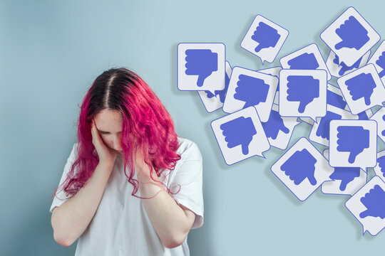 Young woman with pink hair feeling depressed while surrounded by thumbs down symbols, representing online hate, negative comments, bullying, cyberbullying and emotional distress.