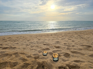 Obraz premium Flip flops on sandy beach with ocean and sunset sky, summer vacation concept
