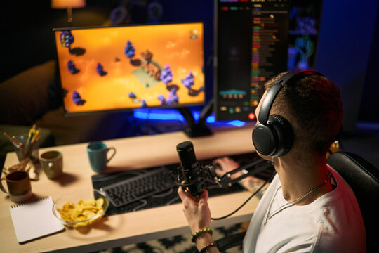 Young adult man streaming gameplay while speaking into microphone at dual monitor desk, creating live gaming content. Ideal for esports marketing, creator economy, online entertainment