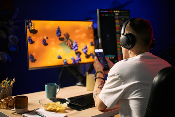 Young adult man streaming gameplay while checking smartphone at dual monitor desk, creating gaming content for online audience. Ideal for esports marketing, creator economy, live stream promotion © DragonImages