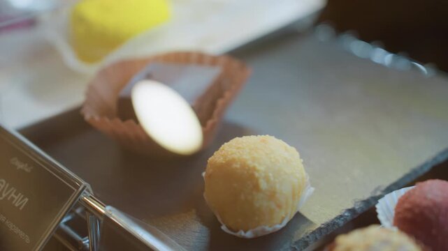 Closeup macarons and truffles display, vibrant yellow macaron and chocolate square in paper cup, glass case reflections and soft natural light, chocolatier arranging samples on boutique counter,