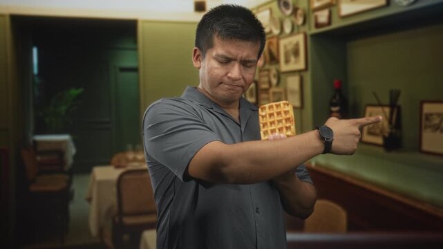 Man holding waffle and pointing finger inside a restaurant building with furrowed brow and crossed arms; confusion.