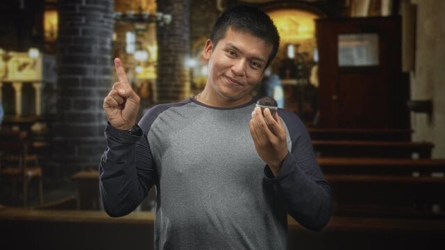 Young hispanic man holding a chocolate muffin in one hand and pointing index finger upward in a church building interior; warm contentment.
