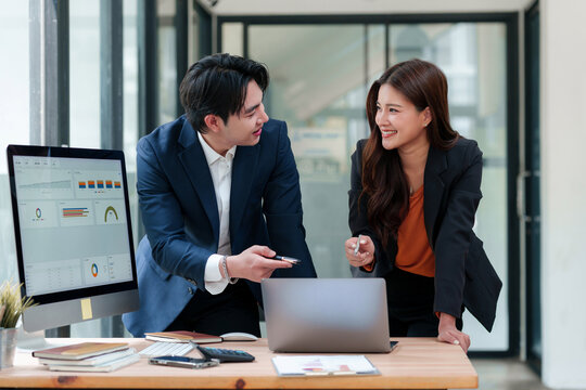 Two professional businesspeople discussing financial data, analyzing reports on a computer screen and laptop in a modern office