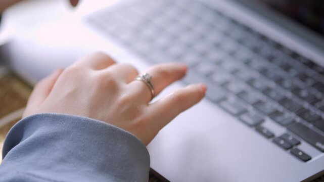 Woman's hands type on laptop keyboard. Fingers press keys. Ring visible on finger. Soft light highlights scene