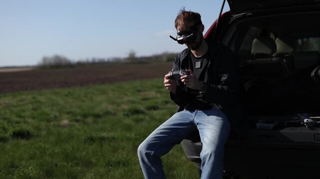 Man sitting in open SUV trunk wearing FPV goggles and holding remote controller while small drone flies in background. Male operator piloting quadcopter in green field on sunny day near his vehicle.