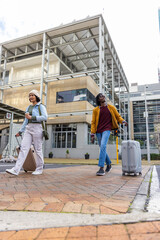 Fototapeta premium Friends walking across brick plaza near glass building, pulling rolling suitcases, holding coffee