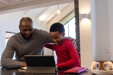 Obraz premium African American father and school-age son leaning over tablet at kitchen counter, smiling together