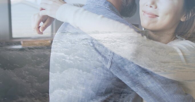Hugging couple standing by window in soft light room, showing wave overlay, sweater and denim shirt