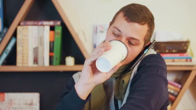 Man on phone sipping coffee, resting chin on hand, bookshelf backdrop and laptop in frame, showing fatigue and distraction during busy remote shift, casual sweater and scarf suggest chilly indoor day