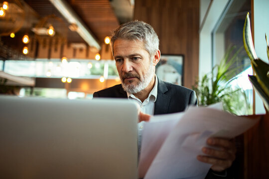 Mature businessman reviewing documents on laptop in cafe