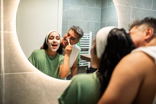 Cheerful adults applying face masks in home bathroom mirror