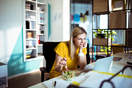 Young woman eating salad while working on laptop in modern office