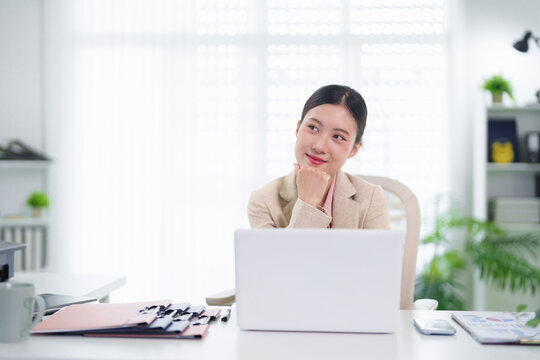 young Asian businesswoman thinking planning strategy while working with laptop computer in modern bright office workspace with natural light and green plants