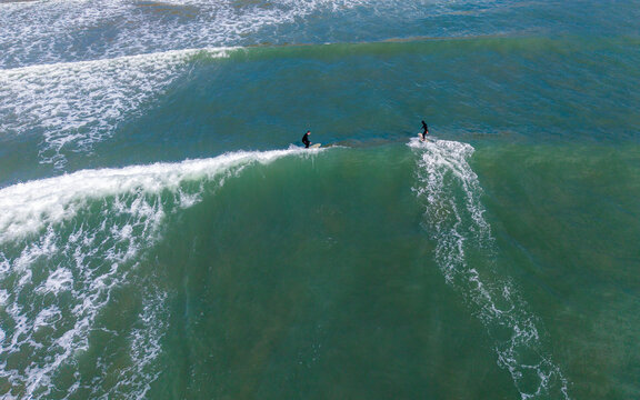 Aerial view of surfers riding the powerful waves, their dark silhouettes contrasting against the turquoise water, Burgh Island, England, United Kingdom.
