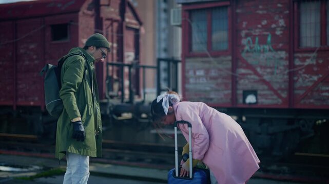 A man and woman, appearing as tourists, stand with luggage in an abandoned railway area. The couple explores a disused train yard as part of their city adventure.