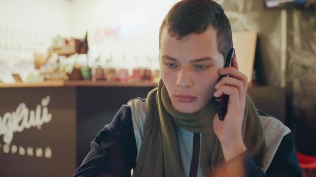 Caucasian man on phone in cafe, covering eyes and leaning forward visible worry, tense gestures, muffled background chatter, chalkboard menu, wool scarf, urgent conversation about news or bills,