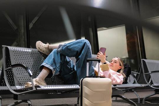 Young female traveler resting on airport bench with suitcase and smartphone, delayed flight and layover concept, night travel, waiting for departure and modern solo travel lifestyle.