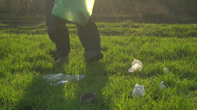 Individual collects plastic waste from grass field using a green trash bag, with various discarded items scattered around in a natural setting