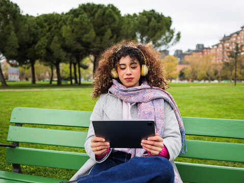 Young woman using digital tablet in park. Curly hair female whit yellow headphones. Whatch movie, tv shows, study, making video call or meeting, conferences