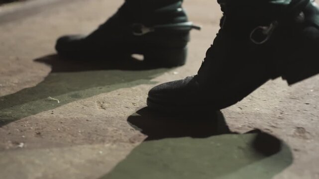 Closeup black boots stepping on pavement, cinematic nighttime street scene with leather buckle catching light, strong shadow, deliberate slow stride, textured concrete and rhythmic footsteps, moody