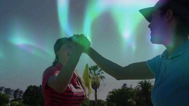 Two women celebrating sinking putt, clasping hands near golf flag, aurora overlay arcing above them