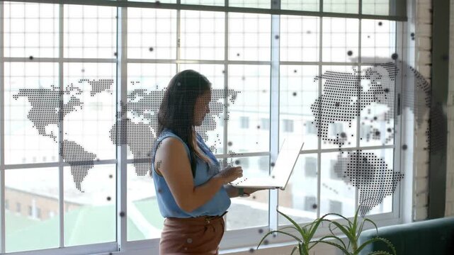Woman standing by grid window tapping laptop and opening screen, dotted world map framing tech work