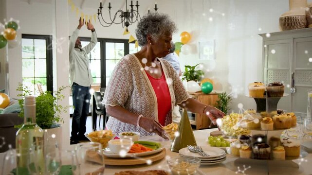 Party prep senior host arranging food on island while family hanging banner, sparkles overlaying