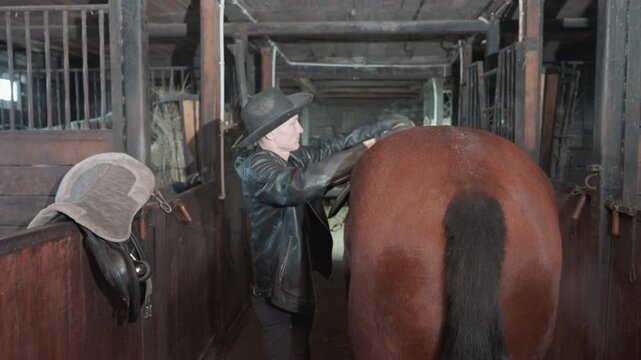 Instructor fitting saddle behind horse in dim stable aisle, hands adjusting girth and strap with saddle set on rail, precise placement and safety check for upcoming riding lesson, focused barn