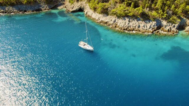 Lone sailboat floating in a bright blue cove surrounded by rocky forested cliffs