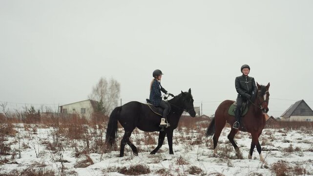 Two white riders standing on snowy field, coach giving verbal guidance during tandem education break, trainee listening beside horse, helmets on, saddles tightened, village houses in distance, quiet