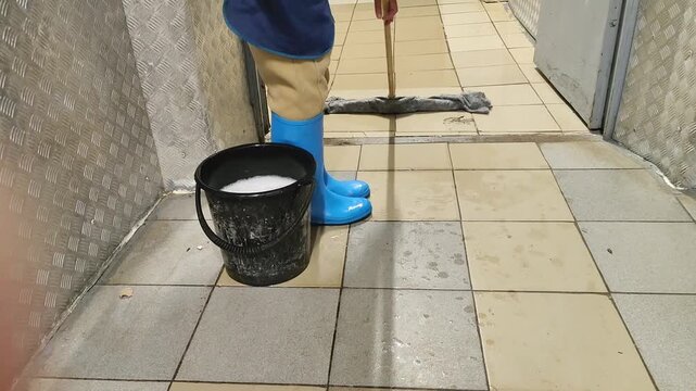 Individual in blue rubber boots mops tiled floor with a wet mop, while a bucket of soapy water sits nearby in a clean indoor corridor setting