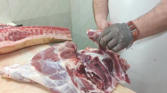 Male butcher in white apron skillfully debones pork leg on wooden table, showcasing precise knife techniques in a clean meat processing area