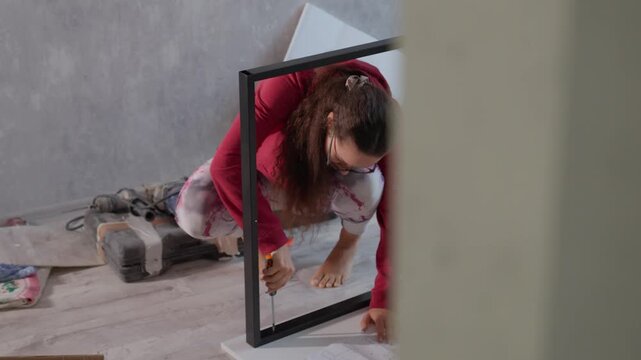 Young Caucasian Woman Kneeling To Insert Hardware While Assembling Furniture, Focused DIYer Uses Screwdriver To Secure Frame To Tabletop, Compact Workspace With Instruction Sheet And Parts Nearby